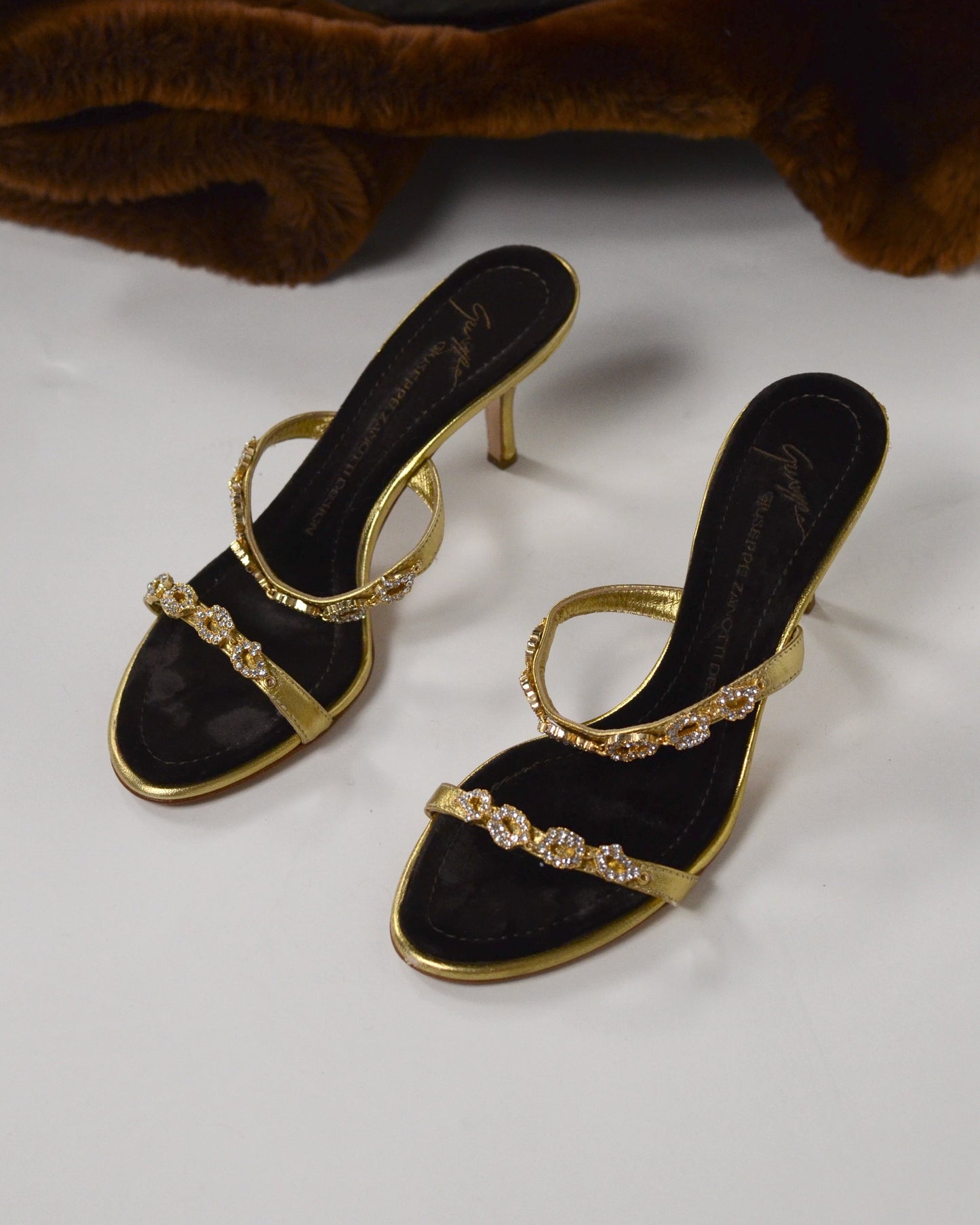 Gold high-heeled  Giuseppe Zanotti sandals with gold straps with  embellishments and brown velvet sole on a white surface.